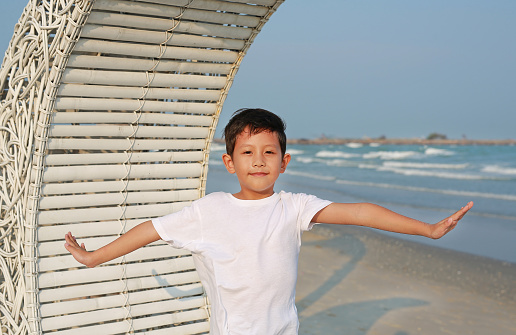 Portrait of smiling Asian boy open arms wide while standing on white bamboo wooden sofa bed with looking camera at the beach.