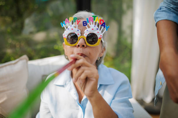 A playful senior woman wearing a birthday hat and glasses blows a whistle toward the camera.