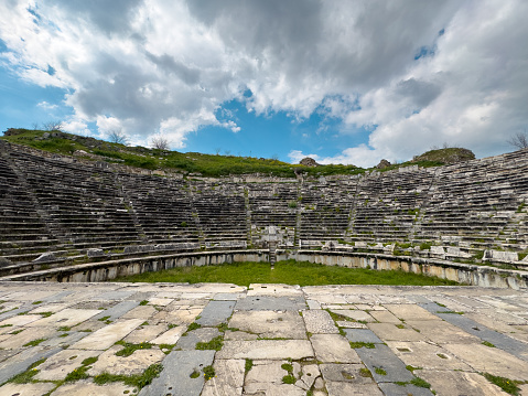 View from the stage of the ancient Roman amphitheater in Aphrodisias, with a dramatic cloudy sky and tiered stone seats.