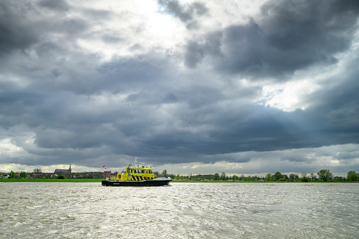 Ship of Rijkswaterstaat sailing on the river IJssel river with high water level on the floodplains of the river IJssel near Zwolle with dark storm clouds above.