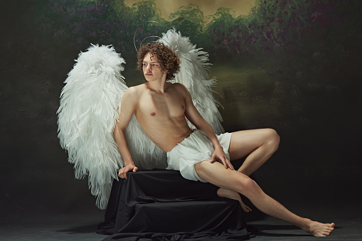 Handsome man with curls and white wings posing radiates sense of peace looks as angel against vintage studio background. Concept of fashion and beauty, modern art and historical fiction fusion. Ad