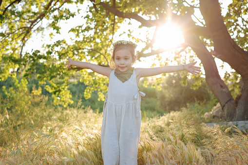 A pretty little girl is picking wildflowers in a lush meadow at sunset. She's alone in a peaceful natural setting, with a forest in the background.