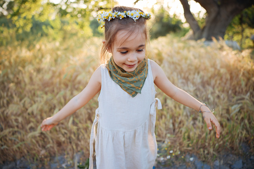 A pretty little girl is picking wildflowers in a lush meadow at sunset. She's alone in a peaceful natural setting, with a forest in the background.