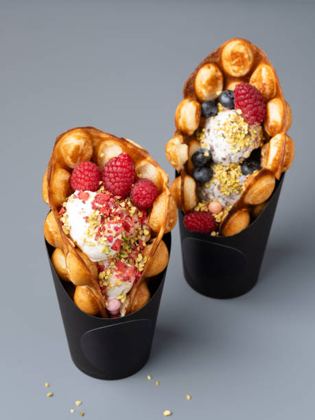 Bubble (Hongkong or egg) waffles stuffed with vanilla ice cream scoops, fresh berries, chopped pistachio and sweet decorations served in black cardboard cups.