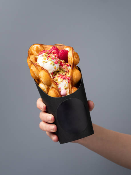 istock Bubble (Hongkong or egg) waffle stuffed with vanilla ice cream scoop, fresh berries, chopped pistachio nuts and sweet decorations served in black cardboard cup in woman's hand. 2150811734