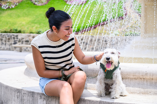 Smiling young woman enjoys a sunny day with her cute dog at a park fountain