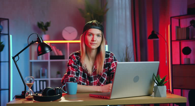Portrait of young woman using laptop and looking at camera at her home workplace in the evening