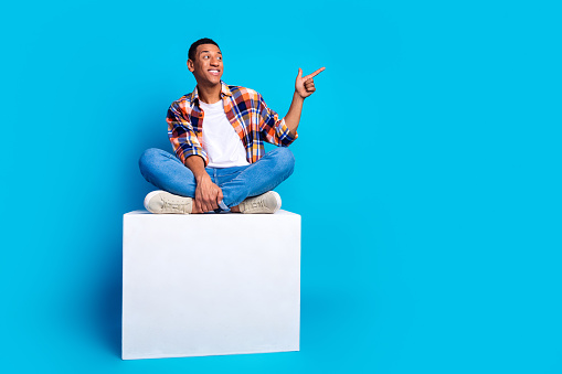 Full length photo of nice young man sit cube indicate finger empty space wear shirt isolated on blue color background.