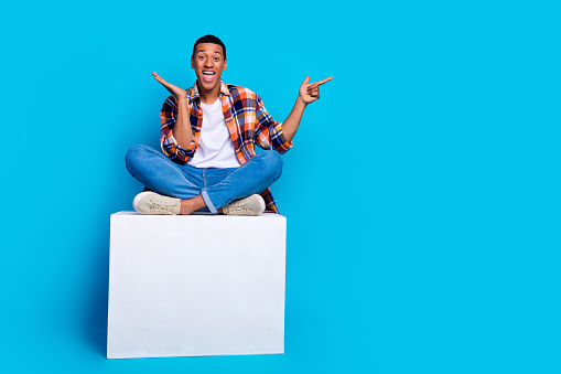Full length photo of young man sit cube indicate finger empty space wear shirt isolated on blue color background.