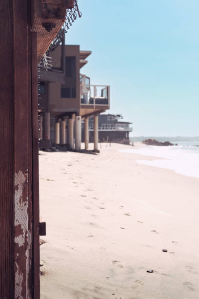 Tranquil beach scene alongside modern beachfront homes. Captured on a clear, sunny day, the image focuses on the sandy shoreline stretching beside a row of contemporary houses with tall supporting pillars. The rustic wooden fence in the foreground adds a textured contrast to the smoothness of the sand, leading the eye towards the ocean's gentle waves in the distance.