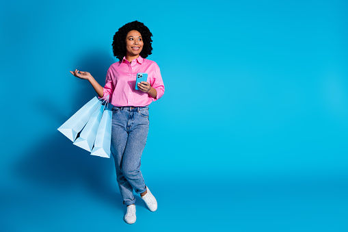 Full length portrait of pretty young woman hold phone bags empty space wear pink shirt isolated on blue color background.