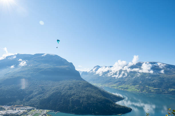 A paraglider soaring above a pristine mountain lake surrounded by majestic peaks and lush green valleys. Brilliant sunlight and clear blue skies enhance the stunning natural scenery.