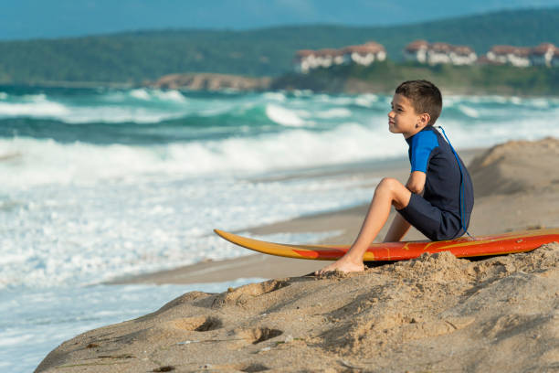 A young boy sits thoughtfully on a surfboard on the sandy beach, gazing at the sea and waves, contemplating surf