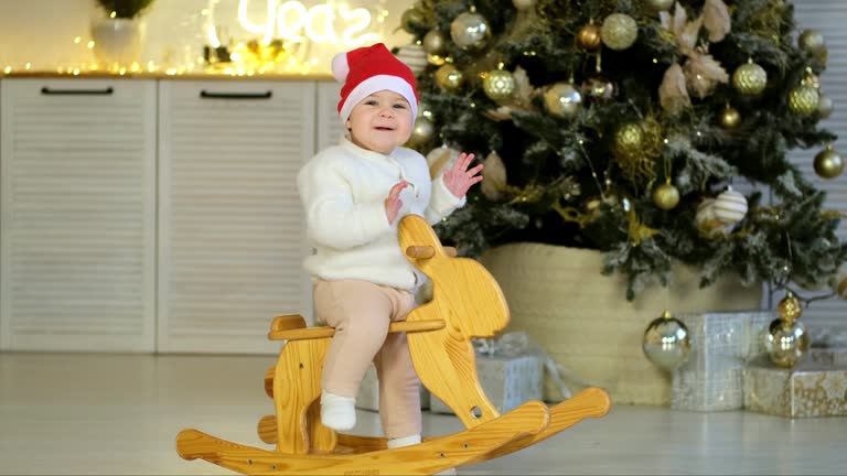 new year at home, the baby is rocking on a rocking horse in a room with a New Year's interior near the Christmas tree, smiling and rejoicing