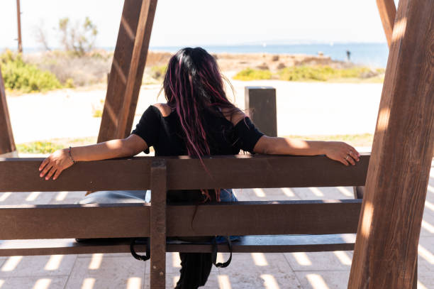 Rear view of a latin woman sitting relaxed on a bench with sea views next to the beach in the shadow