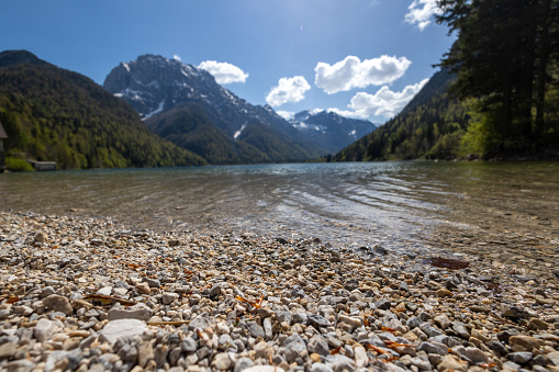Low angle photo of a gravel shore of a calm alpine lake with a view of the mountains in the background and clouds in the sunny sky.
