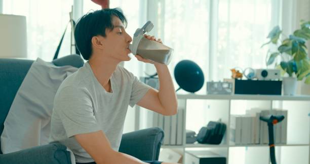 istock Young Asian man drinking nutritious protein shake on sofa in living room at modern home. Diet and healthy food morning routine. 2150865690
