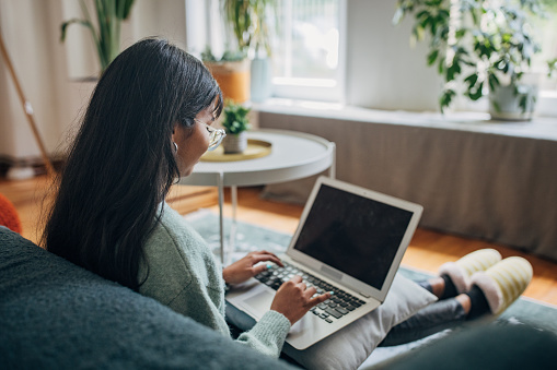 The image captures a focused woman using her laptop in a spacious and bright room, implying productivity and a comfortable work from home setting