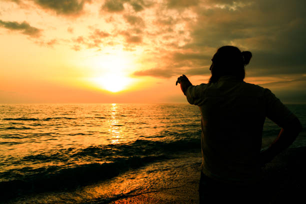 A man stands on the beach looking out at the ocean. The sky is orange and the sun is setting
