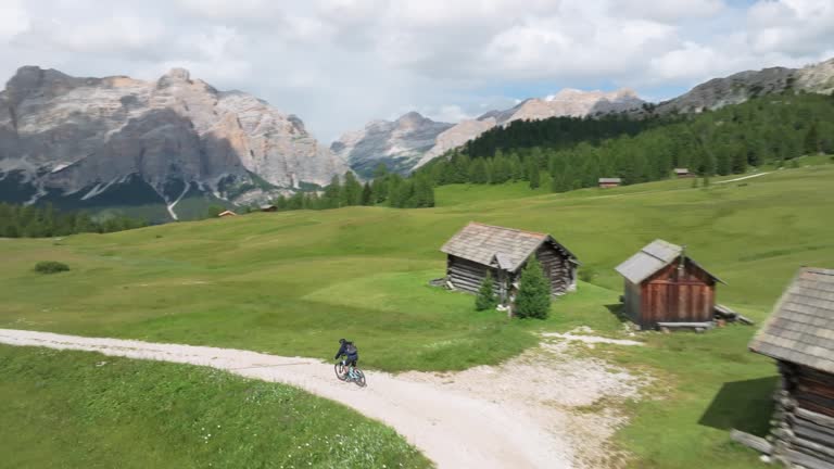 Cyclist rides on winding unpaved road in Pralongia, Italian Dolomites. Aerial cinematic shot captures drone tracking cyclist, passing by wooden cottages. LuPa Creative.