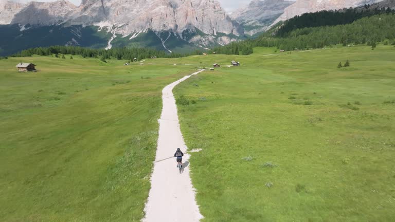 Cyclist maneuvers along winding unpaved road in Pralongia, Italian Dolomites. Aerial cinematic shot showcases drone tracking cyclist's journey through picturesque landscape.
