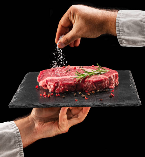Chef is salting or seasoning raw ribeye steak laying on graphite serving board. Black background.