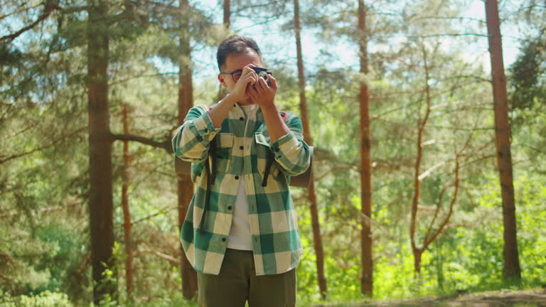 Asian man taking photo in forest, tourist photographer using camera , safe travel and explore, walking hiking tours. Traveling and hiking alone. Summer tourism, wild nature.