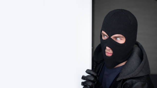 istock An evil thief in mask looks out from behind a white board. Space for text and design. copy space. the surprised bandit looks away 2150889893