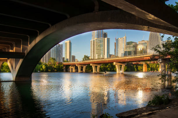 Beneath the South Lamar bridge in Austin, Texas, the tranquil waters of Town Lake and Lady Bird Lake mirror the golden hues of sunset, casting a shimmering glow upon the surrounding high-rise buildings. Against the backdrop of the city skyline, this serene vista harmoniously merges the environment's natural beauty with the modern landscape of urban development.