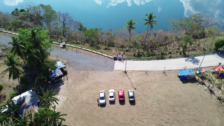 Concrete road constructed in a rural area that runs alongside a serene lake, which is surrounded by dense forests of Lake Tikub, Tiaong, Quezon, Philippines. Drone aerial shot