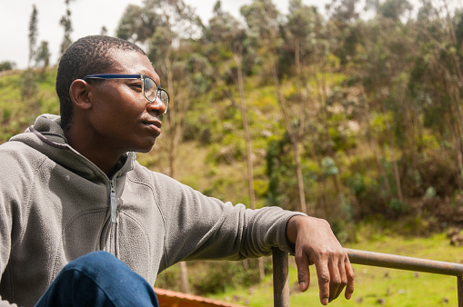 A contemplative young Afro man with glasses leans on a railing outdoors, with green hills in the background.