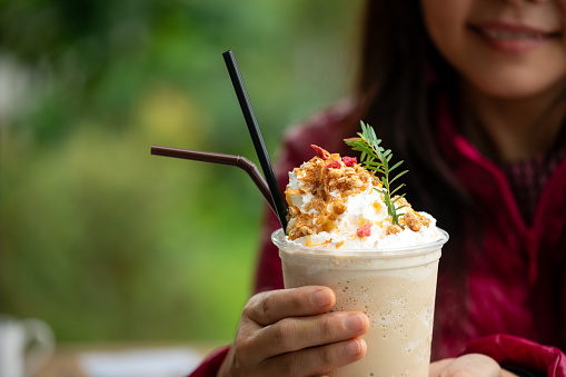 Close-up of woman holding gourmet iced coffee topped with whipped cream and granola, outdoors in lush green setting