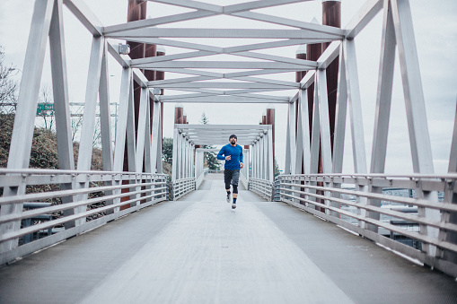 An athletic and healthy man of Filipino descent jogs across a city pedestrian footbridge on a beautiful, chilly Winter day in Portland, Oregon.
