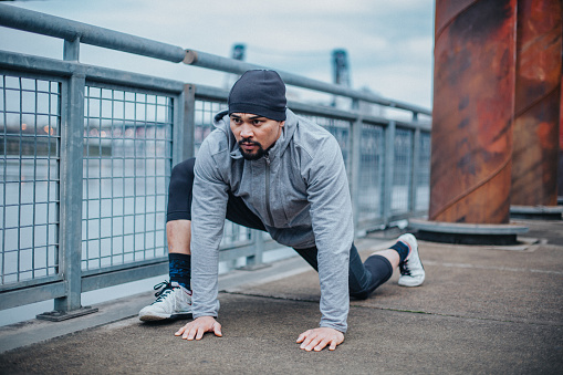 An athletic and healthy man kneels on the floor of a city bridge, stretching his legs before running outside in Oregon on a cold and rainy day.