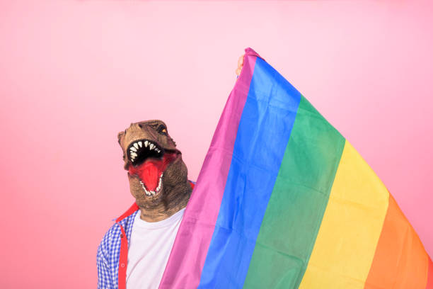 istock Man in T-Rex mask holding a rainbow flag looking at the camera on an isolated pink background 2150913049