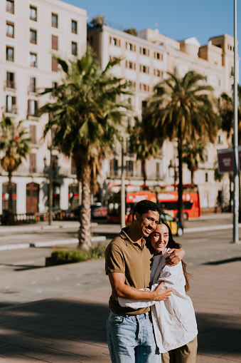 Young couple holds hands and smiles as they walk down a sunlit path in Barcelona, framed by palms