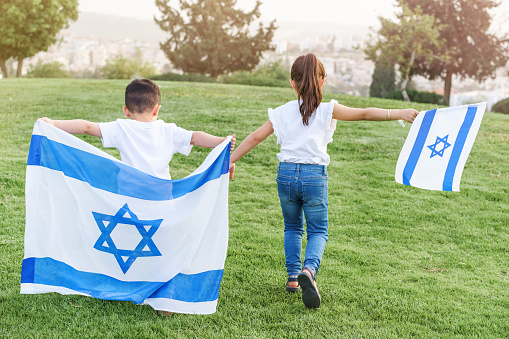 Young children running with Israeli flag. Rear view faceless little girl and boy running with Israel flag up a hill in the park.