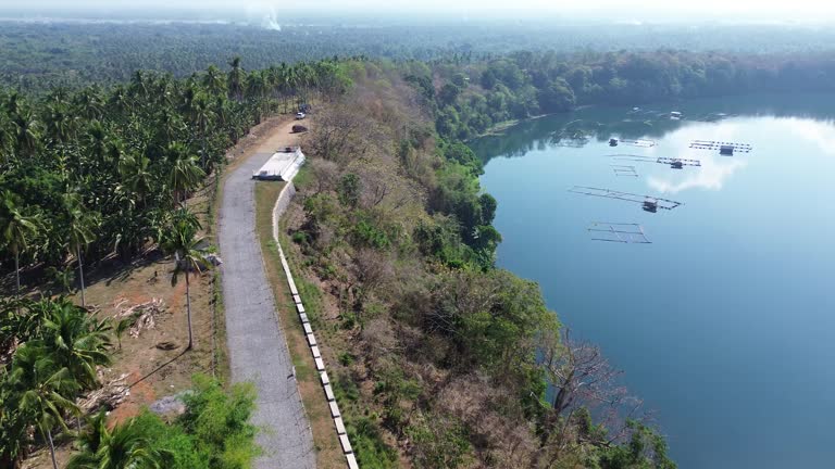 Concrete road constructed in a rural area that runs alongside a serene lake, which is surrounded by dense forests of Lake Tikub, Tiaong, Quezon, Philippines. Drone aerial shot