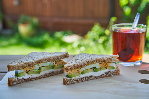 Healty and balanced breakfast served in the garden during sunny summer morning. Sandwich made from wholegrain bread, hard boiled egg, slices of fresh avocado and spreadable fresh cheese with black tea
