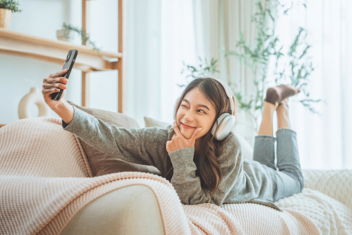 Smiling asian woman with headphones taking selfie with mobile smartphone on couch at home