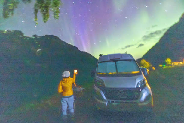 An enthusiastic traveler photographing the night sky beside a camper van. Surrounded by lush greenery and under a canvas of green and purple auroras, this scene shows the perfect blend of adventure and tranquility in the great outdoors.