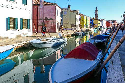 Tourists on Burano Island, on the Venetian lagoon, heading past the islands famous colourful houses and boats on the canal, towards the leaning bell tower of san Martinos church - Venice, Italy