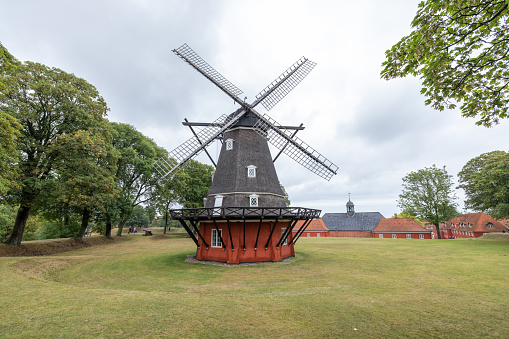 A picturesque traditional Danish windmill stands proudly against a cloudy sky, surrounded by lush green trees and a well-maintained grass field. The historic structure with its dark, tapered body and large, wooden blades is a striking example of Denmark’s cultural heritage and engineering ingenuity, located in a serene rural setting.