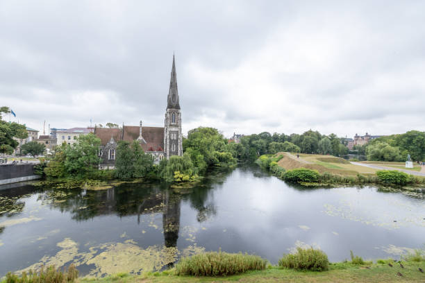 This evocative stock image presents a historic church with an impressive spire, mirrored elegantly in the calm river that flows by it. The overcast sky and verdant riverbanks enhance the peaceful and timeless atmosphere of the scene, making it ideal for themes related to history, serenity, and natural urban landscapes.