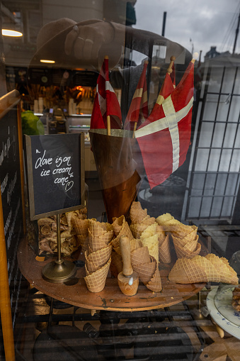 This image captures a quaint ice cream shop display featuring a variety of empty ice cream cones arranged on a circular stand, with a large, swirling ice cream cone model behind them. A Norwegian flag adds a charming touch, reflecting the shop's location or heritage. The display is viewed through a window, with reflections of the street scene subtly visible, adding depth and context to the setting.