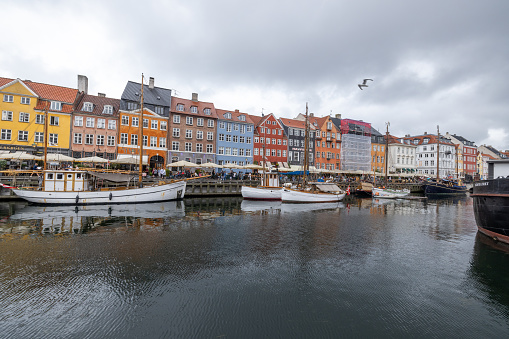 This captivating image features a panoramic view of Nyhavn, the historic and colorful waterfront district in Copenhagen, Denmark. The photograph depicts a cloudy day, with the canal lined with wooden boats and the vibrant facades of old buildings. Tourists and locals enjoy the atmosphere, adding life to the serene waterside setting. The presence of a seagull flying above enhances the maritime feel of this popular Danish destination.