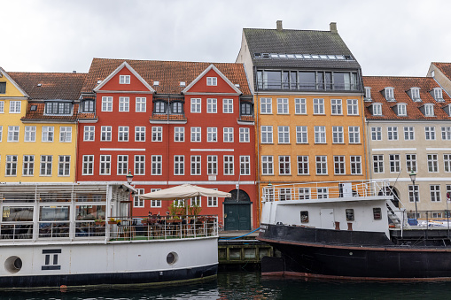 This richly colored image captures the quintessential charm of Nyhavn in Copenhagen, featuring historic ships docked alongside the vibrant, colorful buildings. This part of the canal is known for its lively atmosphere and is a popular spot for dining and socializing, showcasing the blend of Copenhagen’s maritime history with contemporary urban life. The image portrays a typical cloudy day, adding a moody backdrop to the colorful urban landscape.