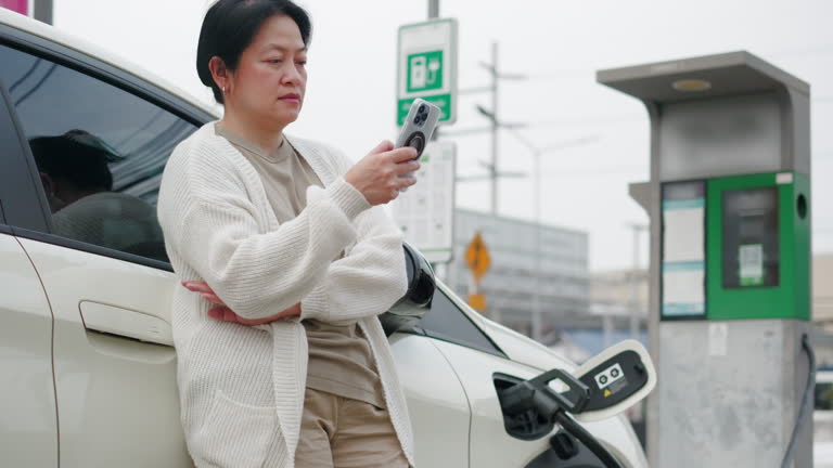 Asian woman use mobile phone connect to see status and waiting when charge  ev car at electronic charging station
