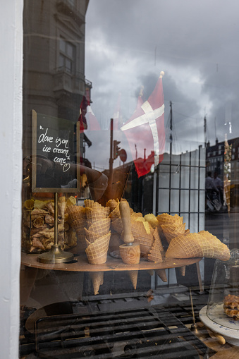 This atmospheric image showcases a charming ice cream shop display through a glass window. The setup includes a variety of waffle cones and a prominent display of a large ice cream model. A Danish flag is featured prominently, suggesting the location or heritage of the shop. The reflection of the cloudy urban skyline in the window adds a moody and intriguing layer to the composition.
