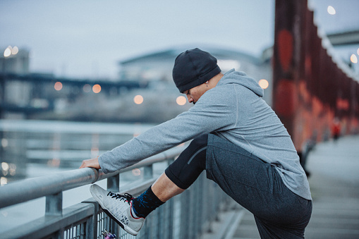 An athletic Filipino man stands on a bridge, stretching his legs before taking a run in a city setting on a rainy, Winter day.